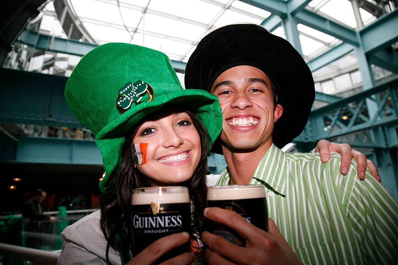 Two people smiling while holding pints of Guinness and wearing St. Patrick's Day hats