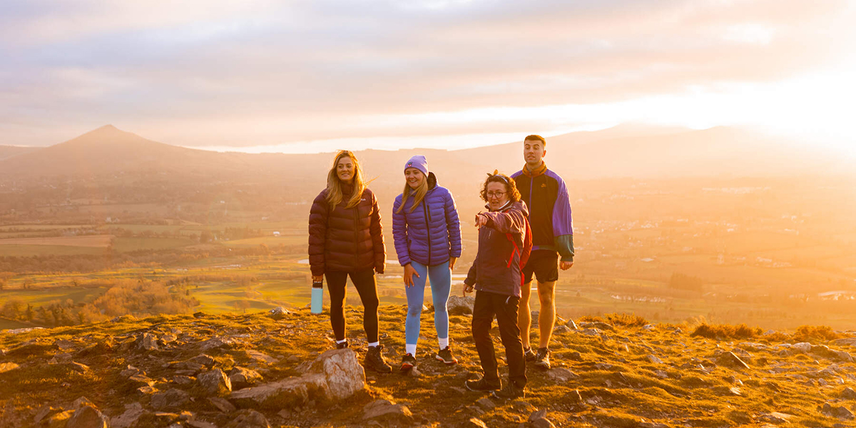 Four people standing on the summit of a hill while the sun is setting and mountains are in background. 