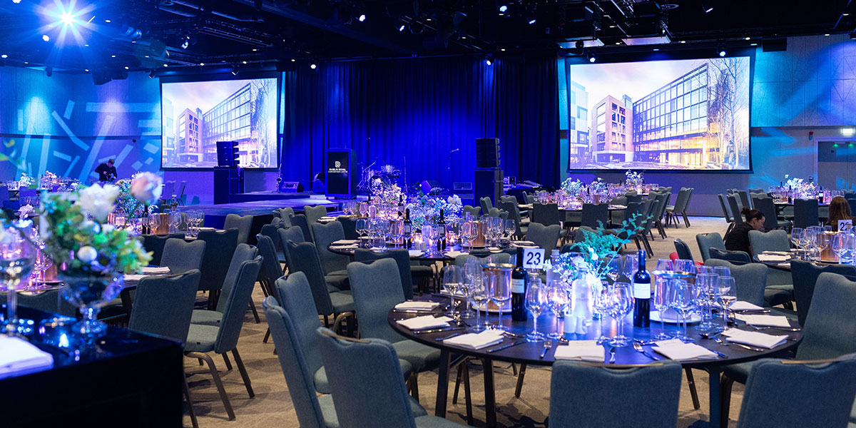 A convention room set up for a formal event, with round tables, floral centrepieces, blue lighting and large projection screens displaying an image of a modern building