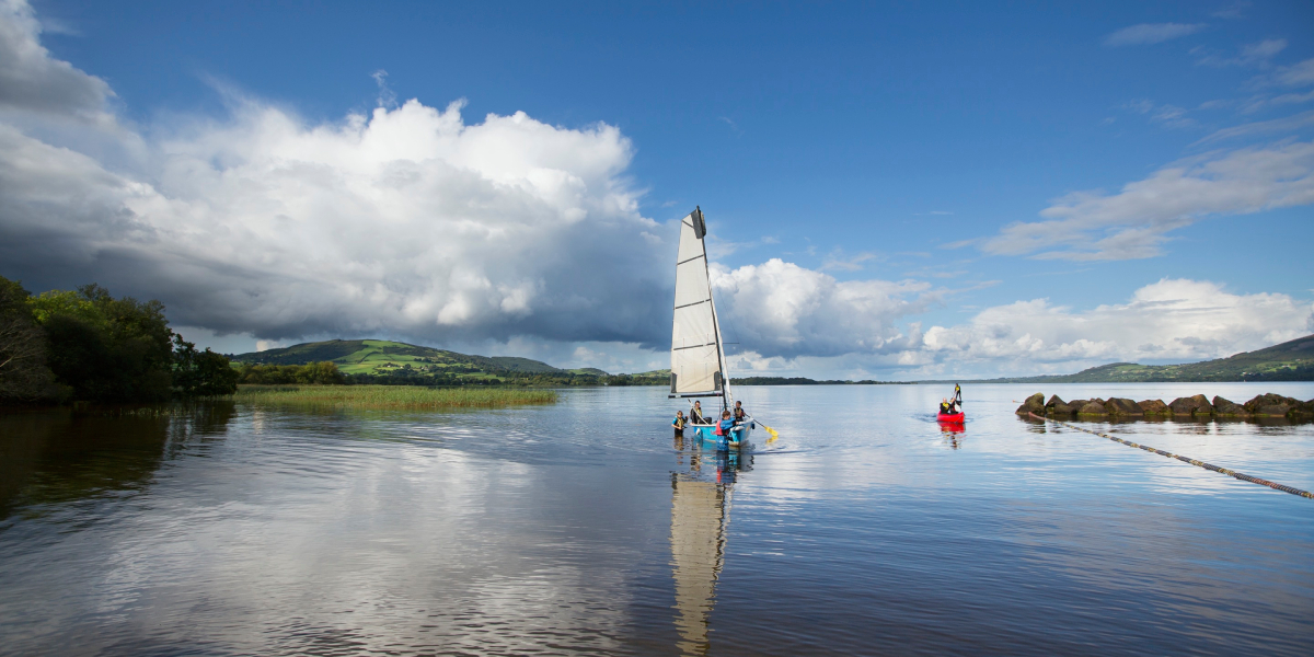Four people sail across Lough Derg in a small boat, surrounded by a clear blue sky and rolling green hills in the background.