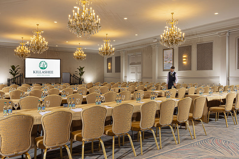 Grand ballroom with rows of gold chairs, chandeliers and a large screen at the front