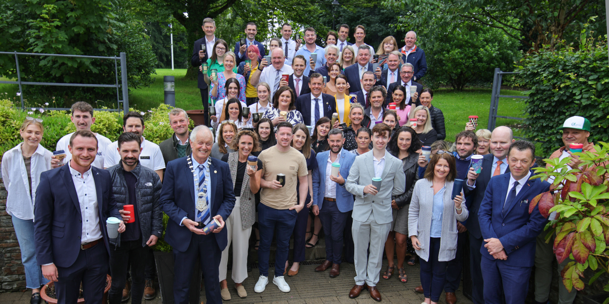 Large group of people posing outside in front of lots of shrubbery holding up reusable coffee cups