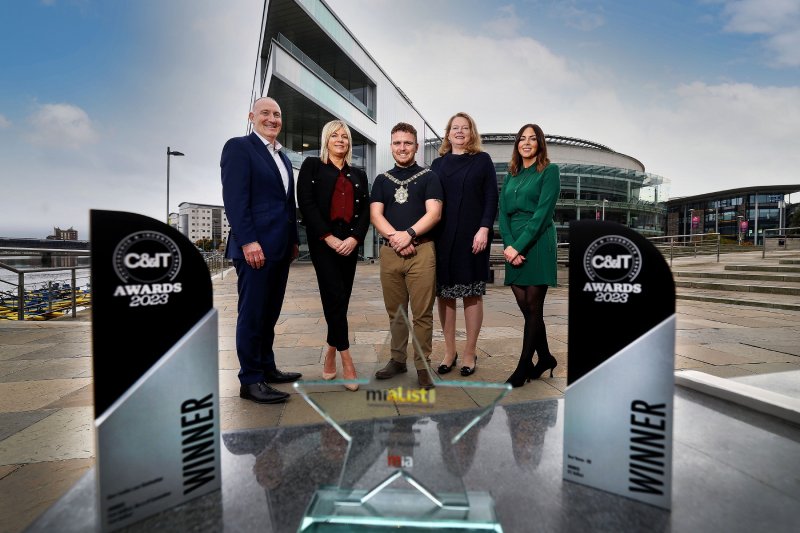 A group of five people smiling and posing outside the ICC Belfast, with a clear blue sky in the background. Three awards are prominently displayed in front of them, showcasing their achievement.